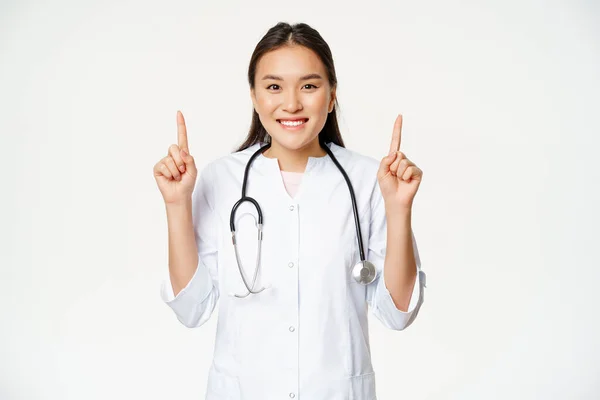 Usmívající se asijské žena lékař, na sobě lékařské roucho, ukazuje prsty nahoru na reklamu, ukazuje promo, poskytují informace o zdravotní péči, bílé pozadí — Stock fotografie