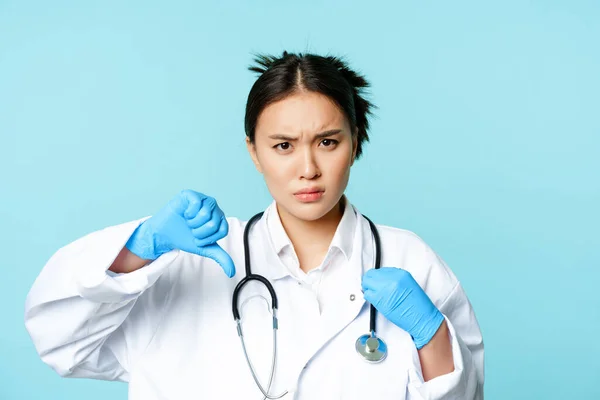 Rozzlobený asijské žena lékař, lékař v uniformě a rukavice ukazuje palce dolů, vrásky obočí rozrušený, neschvaluje smth, modré pozadí — Stock fotografie
