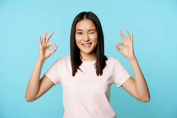Χαμογελαστή κορεάτισσα που κλείνει το μάτι, δείχνει εντάξει σημάδια, συστήνει παρέα ή κατάστημα, στέκεται με t-shirt πάνω από το μπλε φόντο — Φωτογραφία Αρχείου