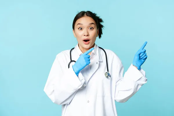 Şok olmuş Asyalı doktor, üniformalı hemşire, sağı işaret ediyor ve şaşırıyor, büyük haber, mavi arka planda duruyor. — Stok fotoğraf
