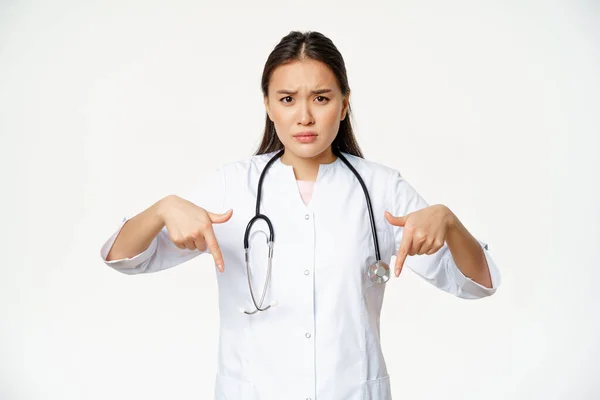 Zklamaný asijský lékař ukazuje prsty dolů, zamračený a vypadá znepokojeně, ukazuje špatný produkt, stojí nad bílým pozadím — Stock fotografie