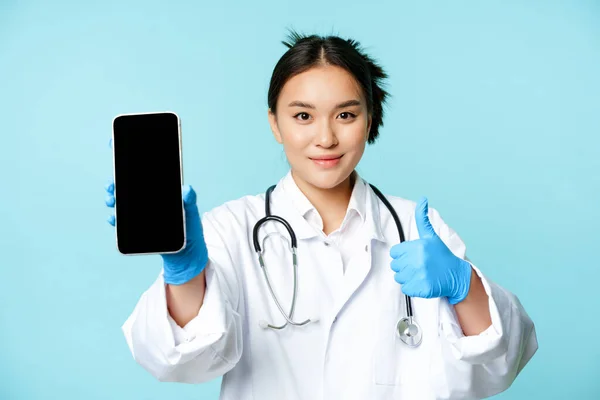Web healthcare, concepto de ayuda en línea. Confiado médico o enfermera asiática, mostrando la pantalla del teléfono inteligente, interfaz de la aplicación y pulgares hacia arriba, recomendando sitio web — Foto de Stock