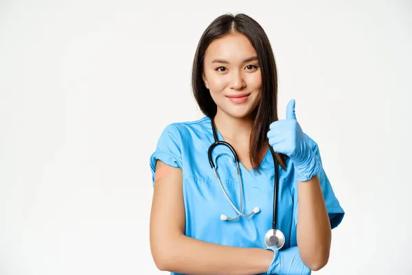 Mosolygó nővér, ázsiai női orvos műtősköpenyben, hüvelykujj fel felirat és beoltott kar orvosi vakolattal, ajánlott vakcinázás covid-19, fehér háttér — Stock Fotó