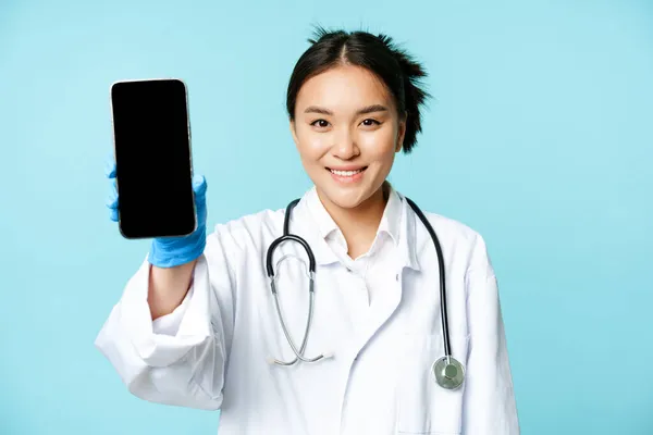 Web healthcare, concepto de ayuda en línea. Sonriendo médico mujer asiática, mostrando interfaz de aplicación de teléfono inteligente, pantalla móvil, de pie en uniforme sobre fondo azul — Foto de Stock