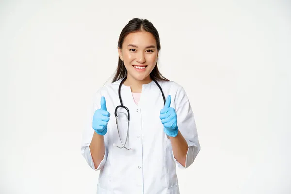 Mosolygó ázsiai orvos, nő orvosi egyenruhában és gumikesztyű mutatja hüvelykujj fel, ajánló smth a beteg, álló felett fehér háttér — Stock Fotó