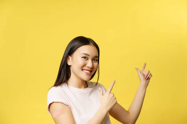 Concepto de vacunación Covid-19. Linda mujer asiática vacunada demostrando pancarta, señalando con los dedos el espacio vacío para el texto promocional, fondo amarillo — Foto de Stock