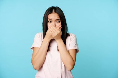Şok olmuş Asyalı kadın ağzını kapatıyor. Çığlıklar atıyor, nefes nefese, korkudan titriyor, dudaklarını kapatıyor, mavi arka planda dikiliyor.