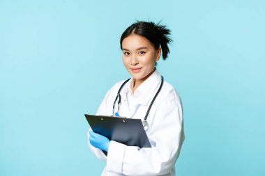 Sağlık ve klinik konsepti. Gülümseyen Koreli doktor, tıp üniformalı kadın doktor, elinde dosya, mavi arka planda duruyor.