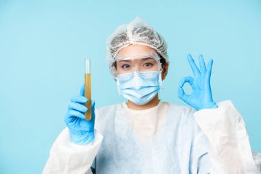 Laboratuvar çalışanı, kişisel koruyucu ekipman hemşiresi, test tüpünde numune gösteriyor ve tamam işareti, test hastası analizi, mavi arka plan.