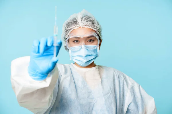 코로나 바이러스와 건강 관리 개념. 아시아 여성 의사 또는 간호사, 감기 치료제를 보여 주는 주사기 개인 보호 장비에 서 있는 푸른 배경 — 스톡 사진