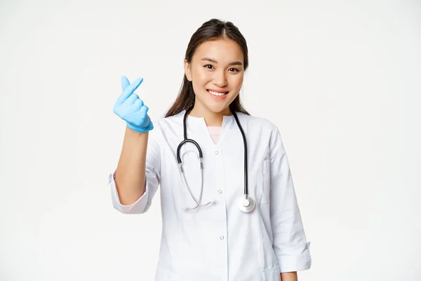 Usmívající se korejský lékař ukazuje prstem srdce gesto v gumových rukavicích, na sobě lékařské uniformy, vypadá šťastně na kameru, bílé pozadí — Stock fotografie