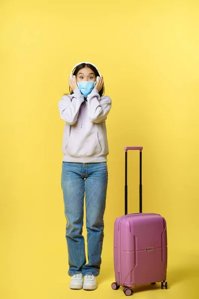 Plná délka záběr šťastně se usmívající asijské turista, dívka ve sluchátkách, poslech hudby na letišti, na sobě lékařské obličejové masky, stojící s kufrem, žluté pozadí — Stock fotografie