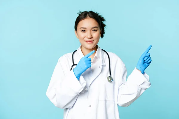 Sorrindo asiático de saúde trabalhador, vestindo luvas estéreis e uniforme, apontando para a direita, demonstrando gráfico, anúncio hospitalar, fundo azul — Fotografia de Stock