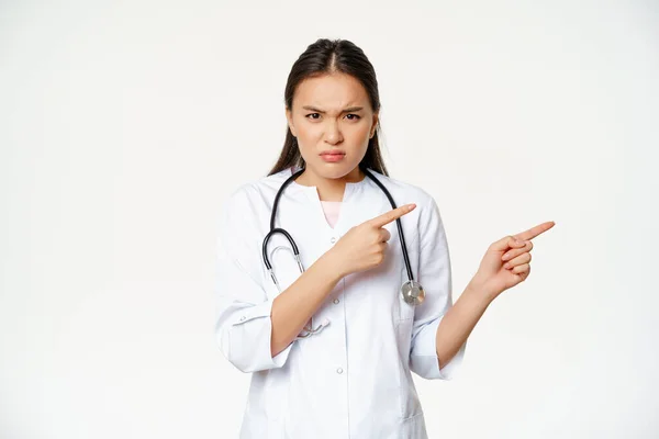 Kızgın kadın doktor, tıbbi cüppeli Asyalı doktor ve steteskop, parmaklarını doğru işaret ediyor ve öfkeli, hayal kırıklığına uğramış, beyaz arka planda. — Stok fotoğraf