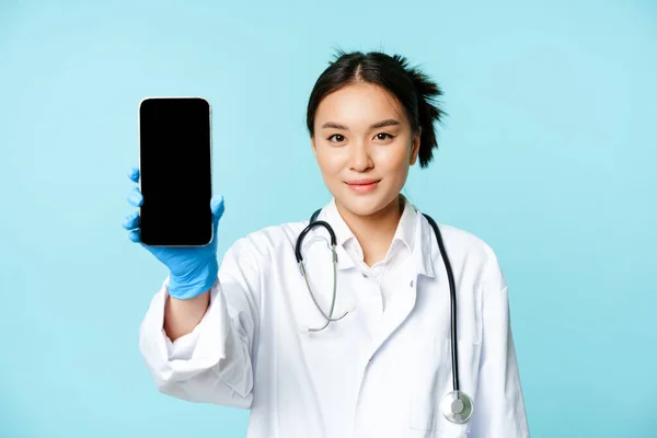 Concepto médico web. Sonriendo médico asiático mostrando la pantalla del teléfono móvil, de pie en uniforme de la clínica, fondo azul — Foto de Stock
