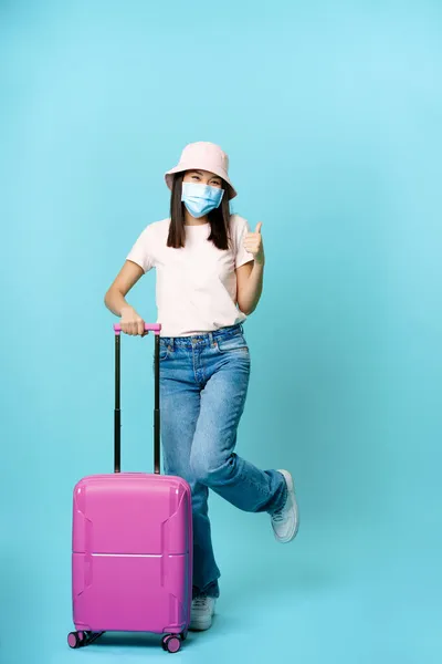 Šťastná korejka v lékařské masce obličeje, palce nahoru, pózování s roztomilou péčí o oblek, turista na dovolené, cestování během covid-19 pandemické koncepce, modré pozadí — Stock fotografie