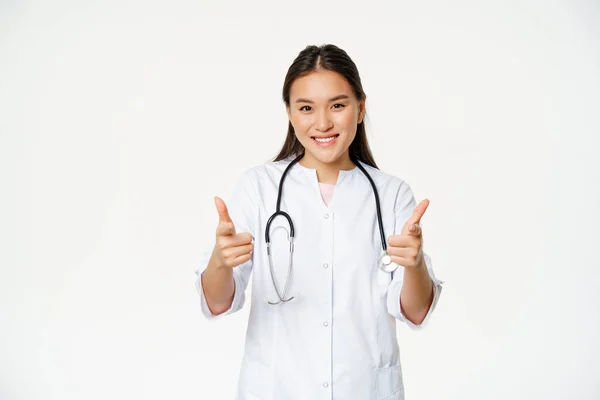Hermosa mujer asiática médico, enfermera en uniforme mostrando pulgares hacia arriba y sonriendo complacido, asegurar paciente, de pie sobre fondo blanco — Foto de Stock