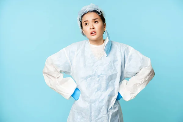 Unavená asijská zdravotní sestra nebo lékař sundat lékařskou masku, pocit únavy, nošení osobních ochranných pomůcek, vzdychání s rukama v pase, modré pozadí — Stock fotografie