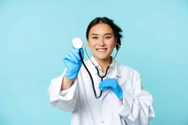 Sağlık ve hasta kontrolü konsepti. Gülümseyen Asyalı kadın doktor, steteskopla muayene eden hemşire, mavi arka planda tıbbi üniforma içinde duruyor. — Stok fotoğraf
