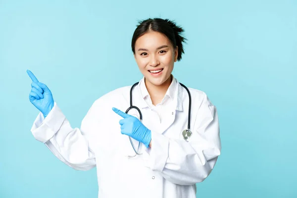Usmívající se šťastná žena lékař nebo zdravotní sestra, ukazuje prstem vlevo, nosí lékařskou uniformu a rukavice, ukazuje nemocniční inzerát, modré pozadí — Stock fotografie