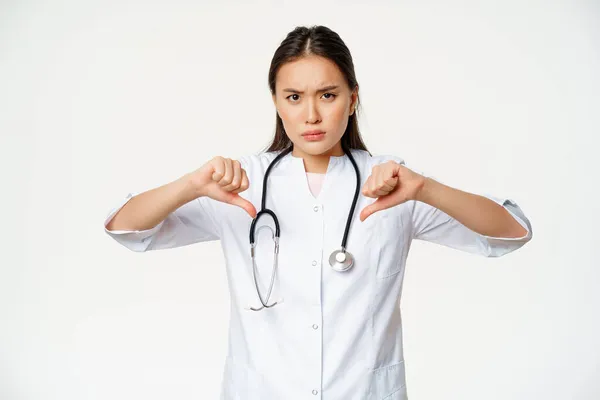 失望した深刻なアジアの女性医師、病院の医師は、親指を示しています不承認で、笑顔悪い嫌い、白い背景の上に立って — ストック写真