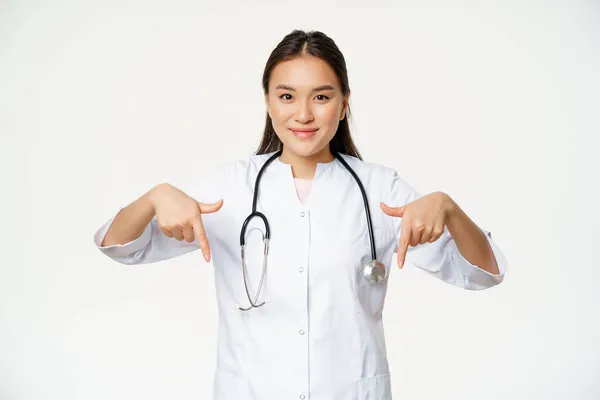หมอสาวเอเชีย ชี้นิ้วลงและยิ้ม สวมกล้องสเตโตสโกป และชุดแพทย์ ยืนอยู่เหนือพื้นหลังสีขาว — ภาพถ่ายสต็อก