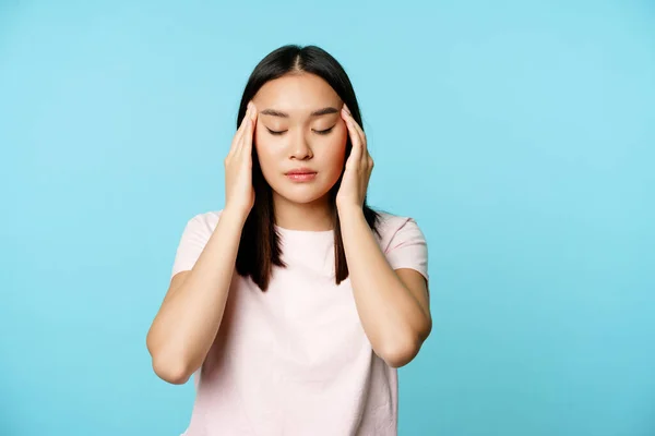 손 과 눈으로 머리를 문지르고, 두 통 으로부터 마사지를 받고, 편두통을 앓고, 푸른 배경 위에 서 있는 아시아 여자 — 스톡 사진