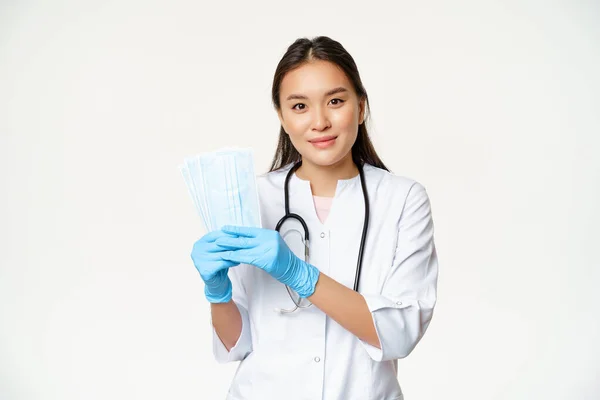 Médico asiático sonriente, médico de familia mostrando mascarillas estériles, previniendo la captura de covid-19, de pie en uniforme sobre fondo blanco Imágenes De Stock Sin Royalties Gratis