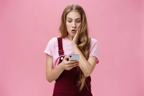 Flicka fick reda på chockerande sanning efter att ha läst meddelande i smartphone trycka arm till kind i överraskning stirrar orolig och mållös på mobilen skärmen reagerar på oväntade nyheter över rosa vägg — Stockfoto