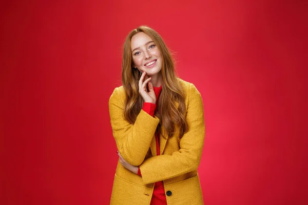 Bien parecido despreocupado y feliz novia pelirroja relajada en abrigo elegante amarillo tocando la cara y la cabeza inclinada como sonriendo con emociones positivas, posando alegre sobre fondo rojo — Foto de Stock