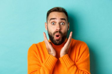 Kameraya bakan, turuncu süveter giyen, turkuaz arka planda duran yakışıklı beyaz adamın yakın çekimi.