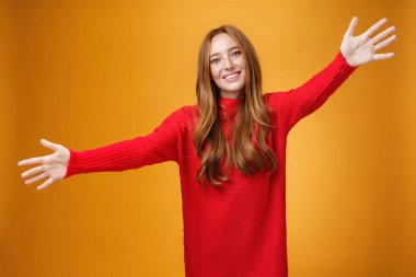 Gel de sarıl bana. Tatlı kadın portresi ve kırmızı örgü elbiseli tatlı kızıl kız kardeş kameraya doğru uzanan ellerini uzatan ve geniş bir gülümsemeyle sıcak karşılayan, turuncu duvarın üzerinde sarılmak isteyen.