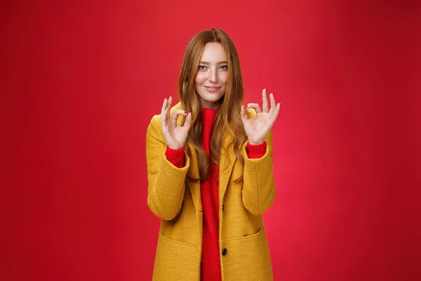 Indoor shot of pleased bright and happy young woman feeling okay and perfect in warm comfortable yellow coat picking right close for cold weather showing ok gesture and smiling delighted over red wall — Stock Photo, Image