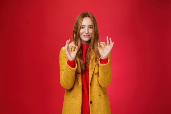 Indoor shot van tevreden heldere en gelukkige jonge vrouw voelt zich goed en perfect in warme comfortabele gele jas plukken recht in de buurt voor koud weer tonen ok gebaar en glimlachen blij over rode muur — Stockfoto