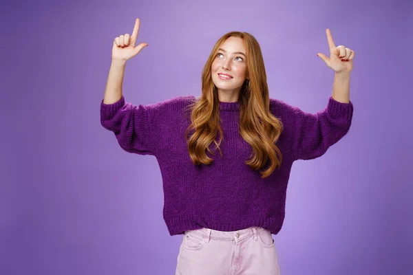 Verträumtes Mädchen mit roten Haaren und Sommersprossen im warmen, gemütlichen lila Pullover, das die Hände hebt und mit fasziniertem und glücklich bezaubertem Gesichtsausdruck nach oben zeigt, lächelnd, wie es neugierige Produkte über violette Wand mag — Stockfoto