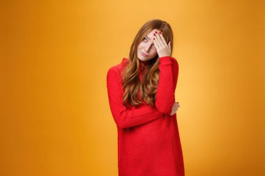 Annoyed and pissed embarrassed woman hiding behind hand making facepalm gesture looking away with irritated humiliated gesture, standing fed up and intense against orange background
