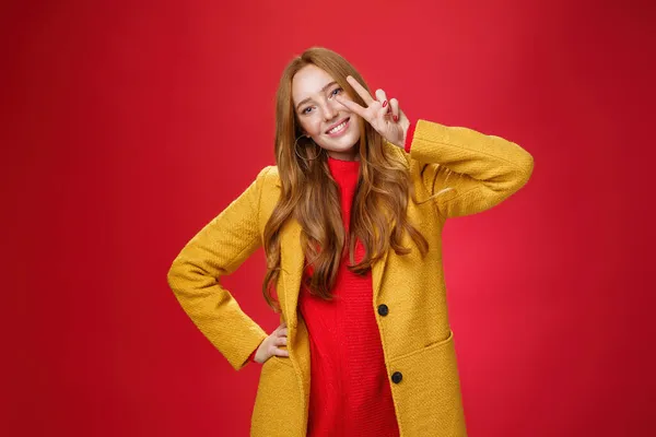 Menina amigável sorrindo feliz, inclinando a cabeça e mostrando paz ou gesto de vitória perto do rosto, segurando a mão no quadril como posando em elegante casaco amarelo elegante e suéter contra o fundo vermelho — Fotografia de Stock