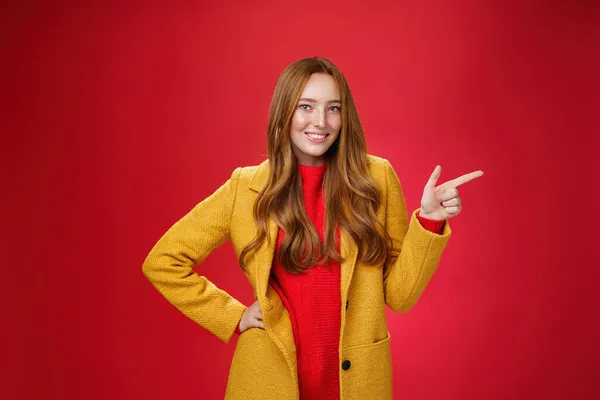 Κομψό και αυτοπεποίθηση ελκυστική κοκκινομάλλα γυναίκα με φακίδες σε κίτρινο εξωτερικό παλτό που δείχνει δεξιά με το πιστόλι δάχτυλο και χαμογελώντας χαρούμενα ως κάμερα, όπως δείχνει δροσερό προώθηση πάνω από τον κόκκινο τοίχο — Φωτογραφία Αρχείου