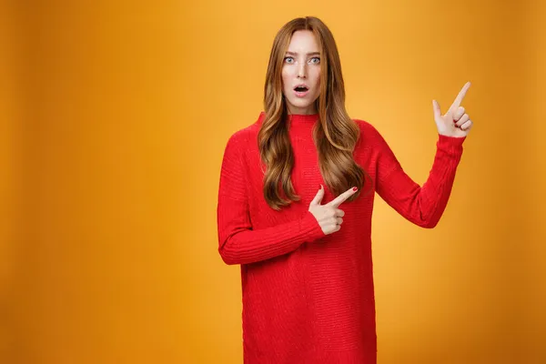 Shocked distressed and confused ginger girl with freckles and blue eyes in red sweater pointing at upper right corner with shook and surprise open mouth, raise eyebrows from stupor over orange wall — Stock Photo, Image