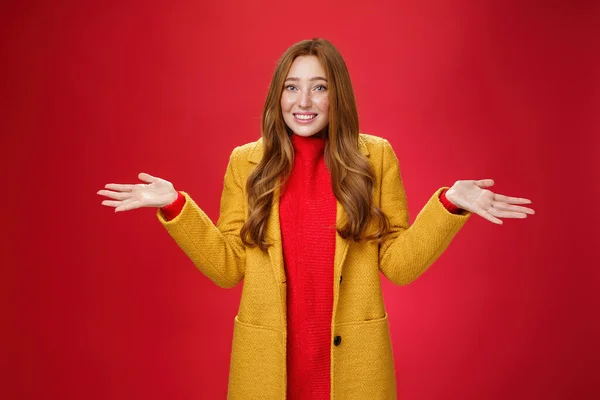 Redhead κορίτσι απολογείται για την καθυστέρηση χαμογελώντας ανόητο και ένοχο με θλιβερό βλέμμα shrugging ως σηκώνοντας τα χέρια πλάγια φορώντας κομψό κίτρινο παλτό πάνω από φόρεμα που θέτουν σε κόκκινο φόντο — Φωτογραφία Αρχείου