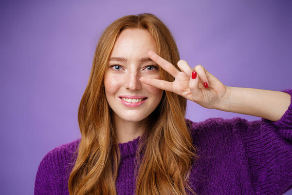 Headshot of friendly and optimsitic happpy redhead female student in purple sweater showing victory or peace gesture over eye like disco dance move smiling broadly over violet background