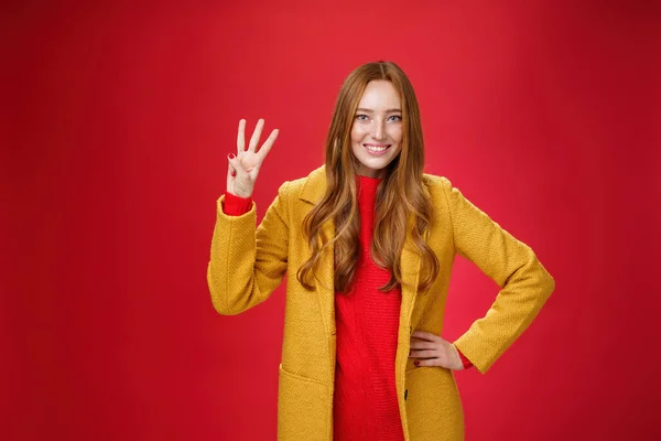 Linda menina gengibre amigável mostrando três razões comprar produto sorrindo amplamente fazendo terceira reserva, olhando encantado e confiante em sua escolha posando contra o fundo vermelho no casaco amarelo — Fotografia de Stock