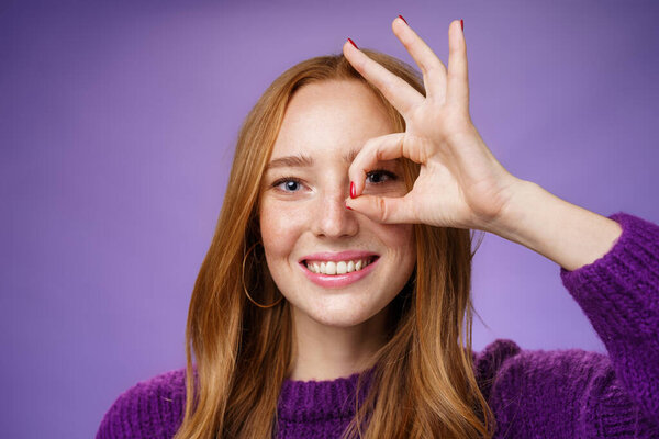 Headshot of optimistic and playful kind cute redhead female with freckles and white perfect smile showing okay or zero sign on eye, grinning as peeking through hole at camera over purple background