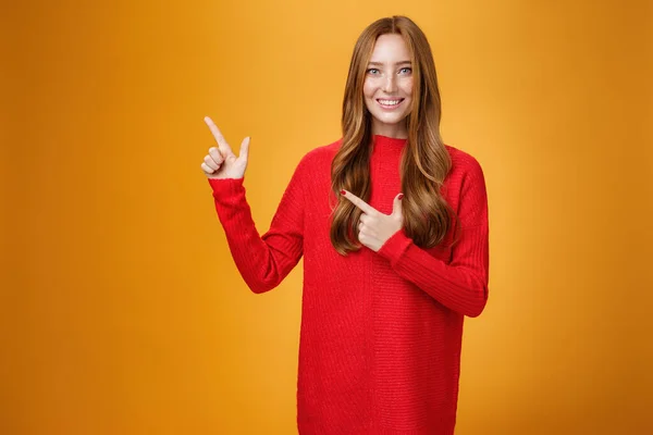 Assirente e disponibile affascinante giovane assitente femminile in abito rosso a maglia con i capelli rossicci sorridente amichevole e piacevole come puntare in alto a sinistra, mostrando cosa interessante sopra parete arancione — Foto Stock