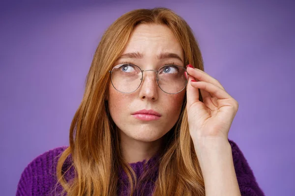 Clumsy and cute young female teacher in glasses with red hair looking unsure and confused looking interested at upper left corner touching rim of glasses thinking or picturing in imagination — Stock Photo, Image