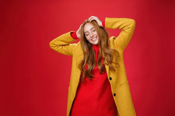Ξέγνοιαστη χαρούμενη κοκκινομάλλα που παρασύρεται με μουσική και καλές δονήσεις κρατώντας τα χέρια στο κεφάλι κλειστά μάτια και χαμογελώντας χαρούμενη και χαλαρή διασκεδάζοντας, χορεύοντας πάνω σε κόκκινο φόντο με κίτρινο παλτό — Φωτογραφία Αρχείου