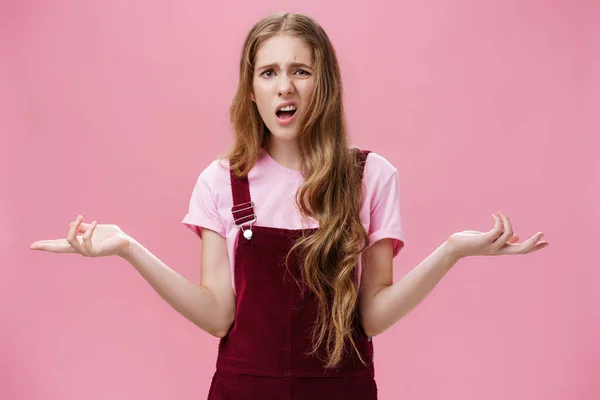 So what, no big deal. Rude and impolite arrogant young girl with high ego in overalls spreading hands sideways in questioned, bothered gesture expressing confusion, feeling pissed with stupid question — стоковое фото