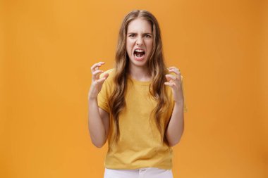 Portrait of irritated cute european female squeezing hands in fists with anger frowning and making hateful face standing annoyed and pissed over orange background with furious look