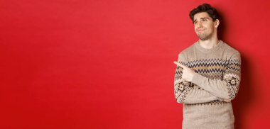 Noel süveteri içinde hayal kırıklığına uğramış yakışıklı bir adam, üzgün bir yüzle bakıyor ve onu işaret ediyor, yeni yıl reklamını yapıyor, kırmızı arka planda dikiliyor.