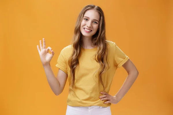 Chica mostrando respuesta positiva con lenguaje corporal. Encantadora joven estudiante de aspecto amigable con un peinado ondulado natural cogida de la mano en la cadera haciendo un gesto bien y sonriendo sobre la pared naranja —  Fotos de Stock